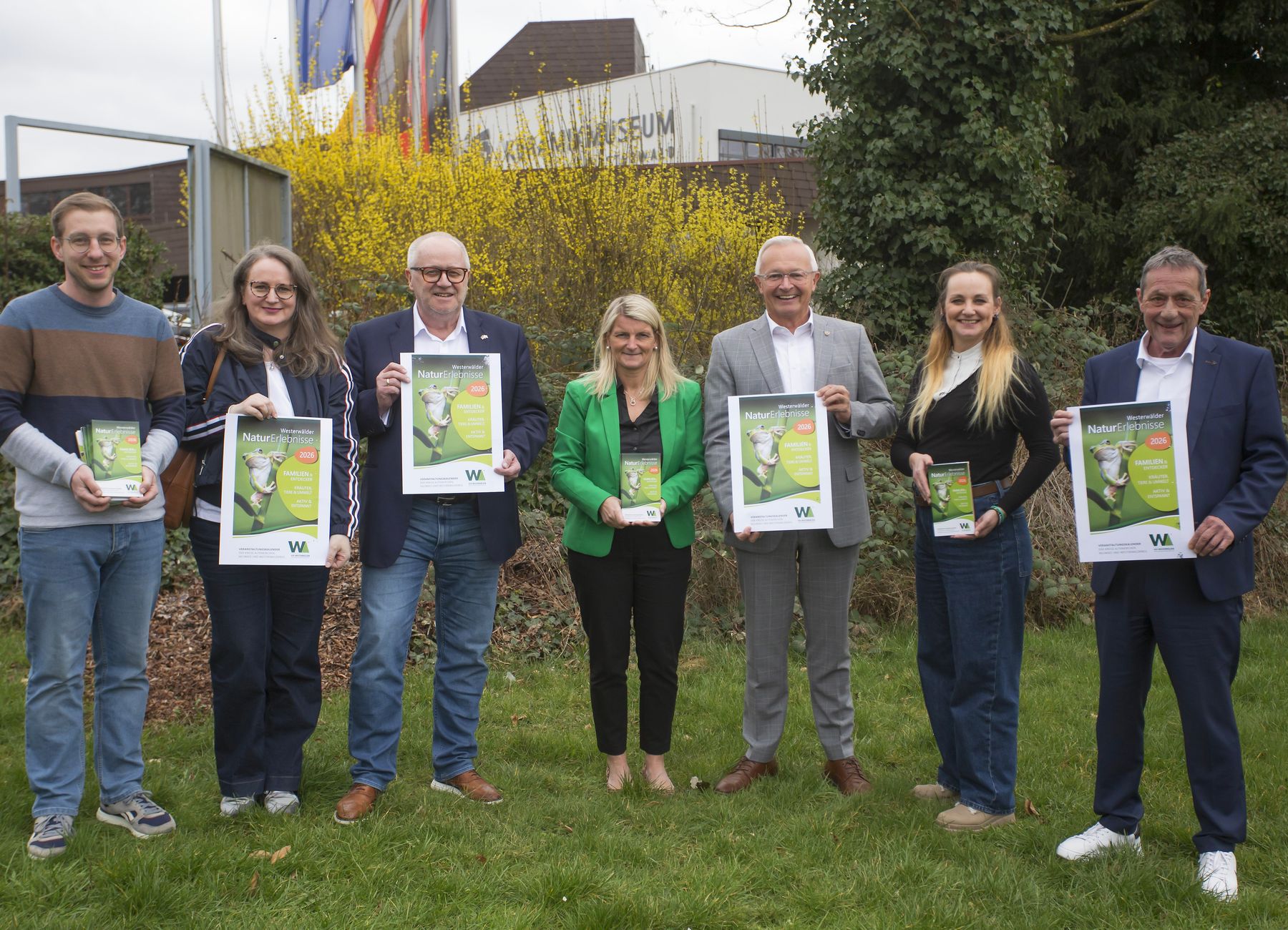 Sieben Erwachsene stehen im Freien im Gras, lächeln und halten Plakate oder Flugblätter in der Hand. Im Hintergrund sind grüne Büsche und ein Gebäude mit Fahnen und einem Schild mit der Aufschrift "MUSEUM" zu sehen.